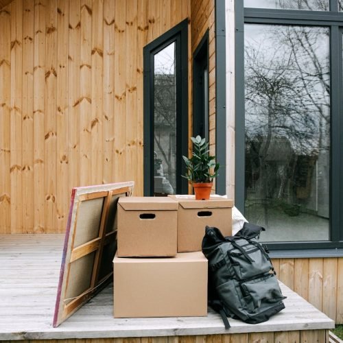 Wooden porch with cardboard boxes and potted plant next to glass windows.